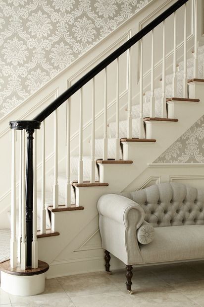 Elegant foyer with silver gray damask wallpaper, wainscoted staircase, glossy black banister and gray tufted settee with caster