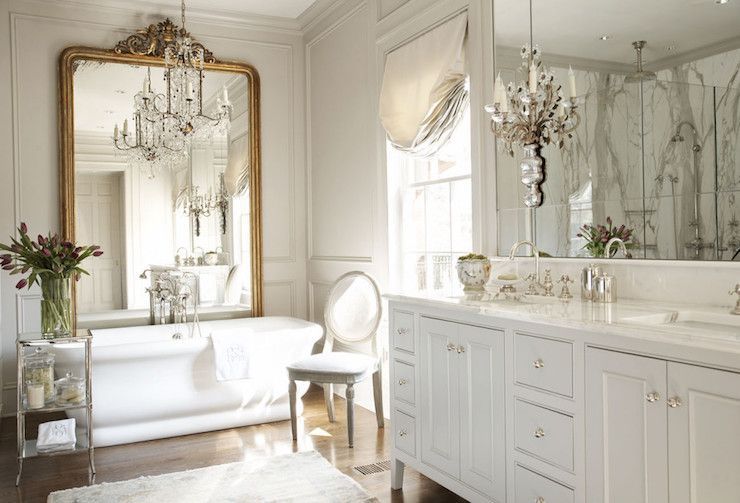 French master bathroom features a white double washstand with feet topped with a white marble framing his and her sinks under mirror illuminated by French sconces. Master bath boasts gray walls accented with gray trim moldings alongside a gilt floor mirror leaning against a wall placed in front of a freestanding tub under a chandelier paired with a polished nickel etagere and an oval back French chair -   22 french decor bathroom
 ideas